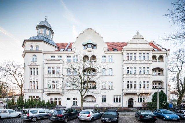 Großes weißes Haus mit rotem Dach, Erkern und Türmchen, davor parkende Autos