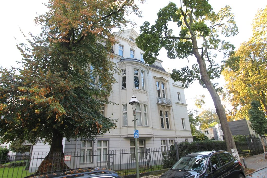 Weißes Haus mit Erker hinter großen Bäumen an einer Straße