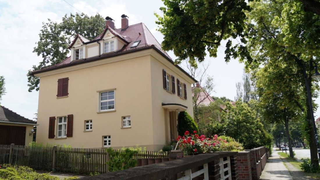 Gelbes Haus mit Dachgauben hinter einem Holzzaun, davor Blumen an einer Straße